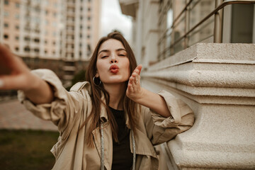 Attractive young charming woman in stylish trench coat blows kiss. Brunette lady in beige outfit...
