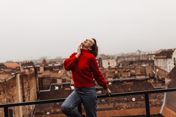 Pretty woman in red sweater and jeans looks up and laughs on balcony. Charming lady poses and smiles on terrace with gorgeous city view.