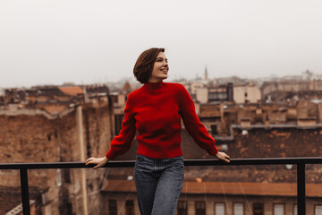 Cool short-haired woman enjoys city view from balcony. Young lady in red sweater and jeans smiles and poses on terrace.