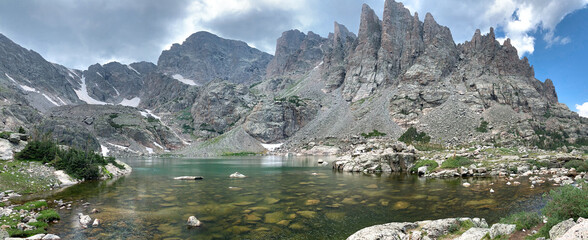 Panoramic view of sky pond in Rocky Mountain National Park in Colorado. Sharp teeth-like peaks can be seen as well as the blue-green water of the pond and patches of snow.