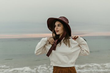 Charming attractive woman in brown pants and white sweatshirt looks away. Young pretty brunette girl in wide-brimmed burgundy hat poses near sea.