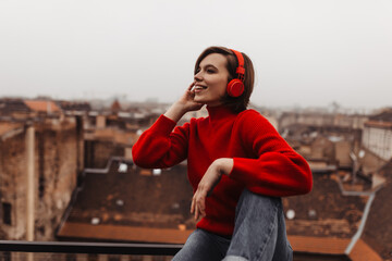 Cool lady in red sweater and jeans listens to music in headphones on balcony. Young woman looks at...