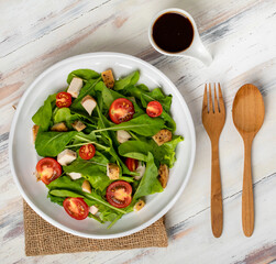 Fresh spinach salad with tomato, small pieces of herb roasted chicken in white ceramic dish on brown sackcloth, together with balsamic vinegar on white wooded table. Concept for healthy