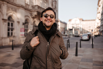 Fototapeta na wymiar Joyful brunet man in sunglasses, scarf and coat smiles widely outdoors. Good-humored guy walks around city.
