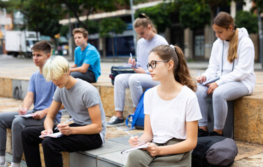 Attentive students write lesson in a notebook sitting on a stone street parapet