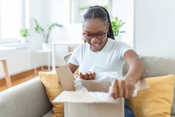 Obraz na płótnie Canvas Happy young woman sit on couch in living room unpack cardboard box buying goods on Internet, smiling excited millennial girl open carton parcel order, shopping online, good delivery concept