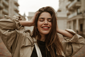 Emotional happy brunette woman smiles sincerely and ruffles hair. Charming woman in beige trench...