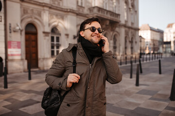 Joyful man in oversized khaki coat and sunglasses speaks on phone. Guy in good mood walks around city.
