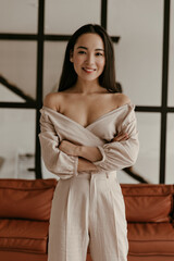 Fototapeta premium Brown-eyed woman in linen stylish pants and beige cropped blouse crosses arms, looks into camera, smiles sincerely and stands in living room.