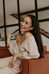 Fototapeta premium Pretty brunette Asian woman in linen blouse and beige pants plays hair, smiles sincerely and sits on brown sofa in living room.
