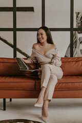 Obraz premium Elegant happy woman in linen pants and beige blouse sits on brown sofa and reads magazine. Charming girl in stylish outfit smiles in living room.