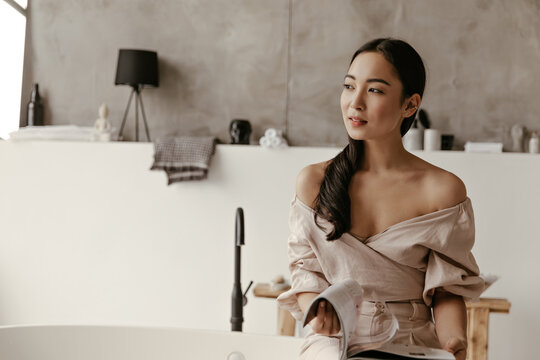 Attractive woman in linen beige blouse and pants looks away, holds fashion magazine and sits on bath in white bathroom.