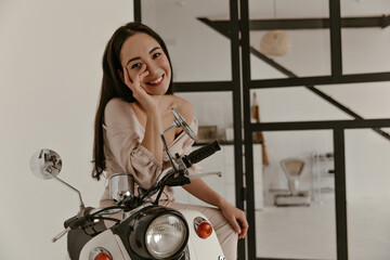 Fototapeta na wymiar Brown-eyed brunette woman in trendy linen blouse and beige pants smiles, looks into camera and sits on moped in white cozy apartment.