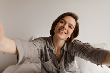 Fototapeta premium Short-haired woman in grey shirt smiles widely, takes selfie in bedroom. Charming lady in pajamas sits on white bed.