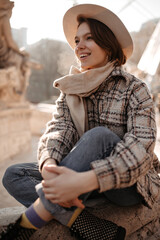 Pretty short-haired woman in stylish checkered coat and jeans smiles widely outside. Young lady sits near fountain.