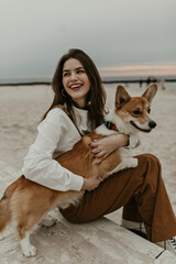 Joyful woman hugs corgi and sits on sand at beach. Attractive cheerful lady on good mood poses with dog near sea.