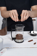 Cropped shot: lady in black is making coffee and breaking cinnamon stick above french press with ground coffee. The press pot is made as glass bulb in stainless body with black handle. 