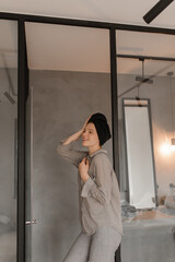 Charming woman in grey shirt, pants and black towel on head smiles. Elegant pretty lady poses in good mood in bathroom.