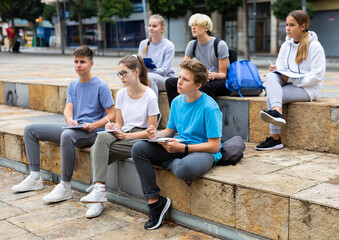 Group of teen students sitting with workbooks in schoolyard in warm autumn day.