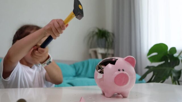 Little child girl is crashing piggy bank with hammer at home in slow motion. High quality FullHD footage