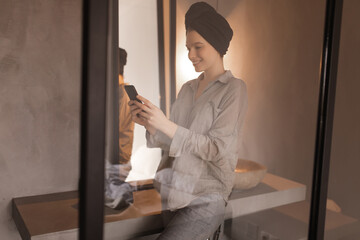 Cool woman with towel on head holds phone. Charming lady in grey shirt smiles and poses near mirror in bathroom.