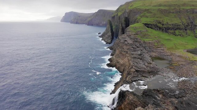 Aerial view of rock cliff in Faroe Islands. Blue sea waves, rough sea on cloudy journey. slow motion