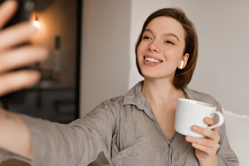 Joyful delighted woman in grey linen shirt smiles, takes selfie and holds white coffee cup. Cool girl poses in bedroom.