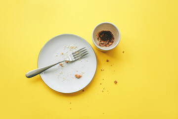 Empty plate with fork and crumbs after eating and cup with coffee grounds on yellow background. Concept of the end of the party or celebration.