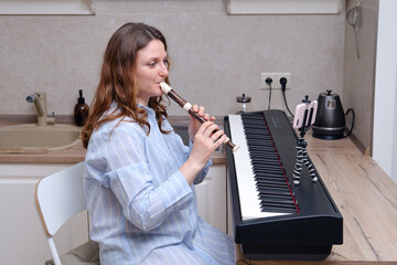 A female musician plays a block flute while sitting at an electric piano in a home kitchen