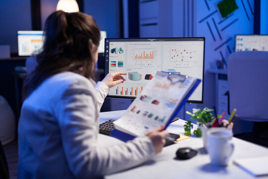 Freelancer woman comparing graphics from clipboard with grafics from computer at business office. Tired focused employee using modern technology network wireless working on project solution