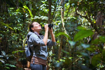 Smiling woman tourist watching birds with binoculars in the rain forest - 447433967