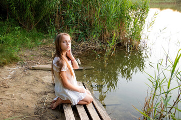 A girl in a white shirt is sitting on the shore of a lake at sunset, illuminated by the evening sun.