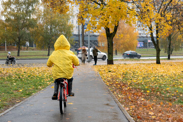 Child rides bicycle in an autumn park during the rain. Boy in yellow raincoat rides a bicycle around the city. Back view