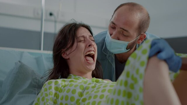 Pregnant young woman giving birth to child feeling tired and screaming in hospital ward bed. Caucasian patient pushing for baby delivery, receiving help from multi ethnic maternity team