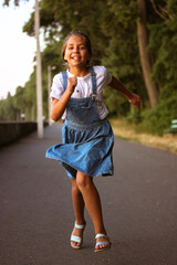 A happy teenage mestizo girl in a denim sundress running along a promenade outdoors. A beautifully tanned 10-12 year old female kid, child on a move. End of summer, back to school. Summer holidays.