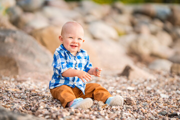 A little baby boy in a blue shirt plays with stones and shells on the beach