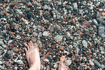 Female feet in sea water on a pebble shore top view in summer, many pebbles, copy space, relaxation in nature, swimming in sea