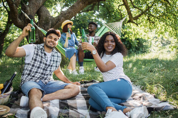 Indian man and african woman relaxing on played on grass with beer in hands while another couple sitting in hammock and chatting on background. Happy friends enjoying picnic outdoors.