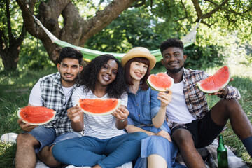 Four cheerful multiracial friends sitting at green garden with slices of sweet watermelon in hands. Young people having picnic on fresh air with tasty food.