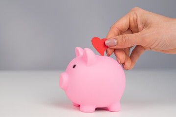 Faceless caucasian woman folds a small heart into a pink piggy bank on a white background. Love saving concept