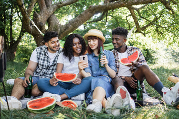 Group of four multiracial friends using modern smartphone, eating watermelon and drinking beer. Happy young people spending free time on nature. Summer picnic at garden.
