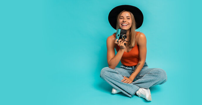 Lovely blond woman in  stylish summer outfit holding retro camera , sitting on floor in studio on blue background. Vacation mood.