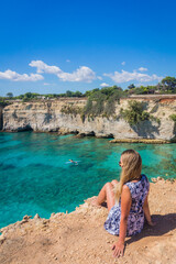 Young woman with long blond hair in a dress relaxing on a cliff and looking at kayak and beautiful turquoise water of Torre Sant Andrea, province of Lecce, Apulia, Italy
