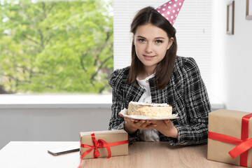 birthday in the office. Against the background of a window with blinds, a young beautiful lady is sitting holding a cake in her hands, smiling and looking at the camera. - Powered by Adobe