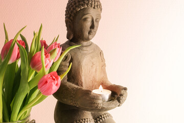 Small buddha sculpture made from ceramic with a burning candle and pink tulips against a light...