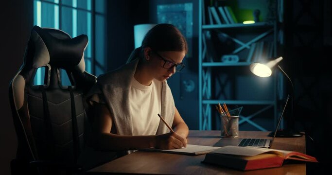 Smart teenage girl sitting in dark room, studying late at night. Bright schoolgirl writing essay in notebook, focused on homework. College student preparing for exam.