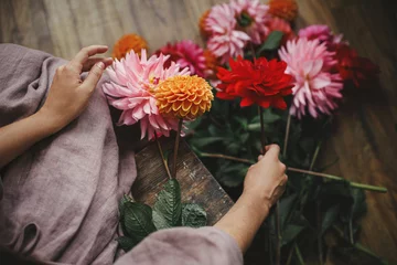 Zelfklevend Fotobehang Woman holding dahlias flowers and sitting on wooden rustic bench, view above. Atmospheric moody image. Florist in linen dress arranging autumn flowers bouquet in countryside. Slow life © sonyachny