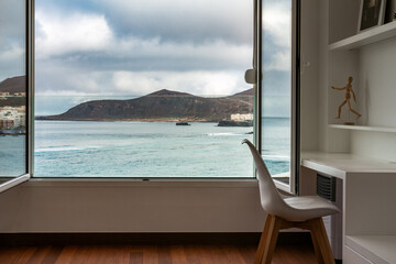Atlantic ocean and landscape view through open window from inside home or hotel room with working table and chair. Work remote from home office in beach apartment. Gran Canaria, Canary Islands, Spain. - obrazy, fototapety, plakaty