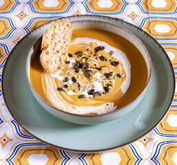 Flat lay photography of a vegan pumpkin cream soup and bread with selective focus. Vegetables recipe. Top view of an autumn puree of seasonal products with a retro pattern fabric tablecloth.