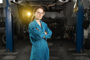 A young woman in work clothes, an apprentice stands next to the car and is proud and happy in the garage with protective goggles. - obrazy, fototapety, plakaty